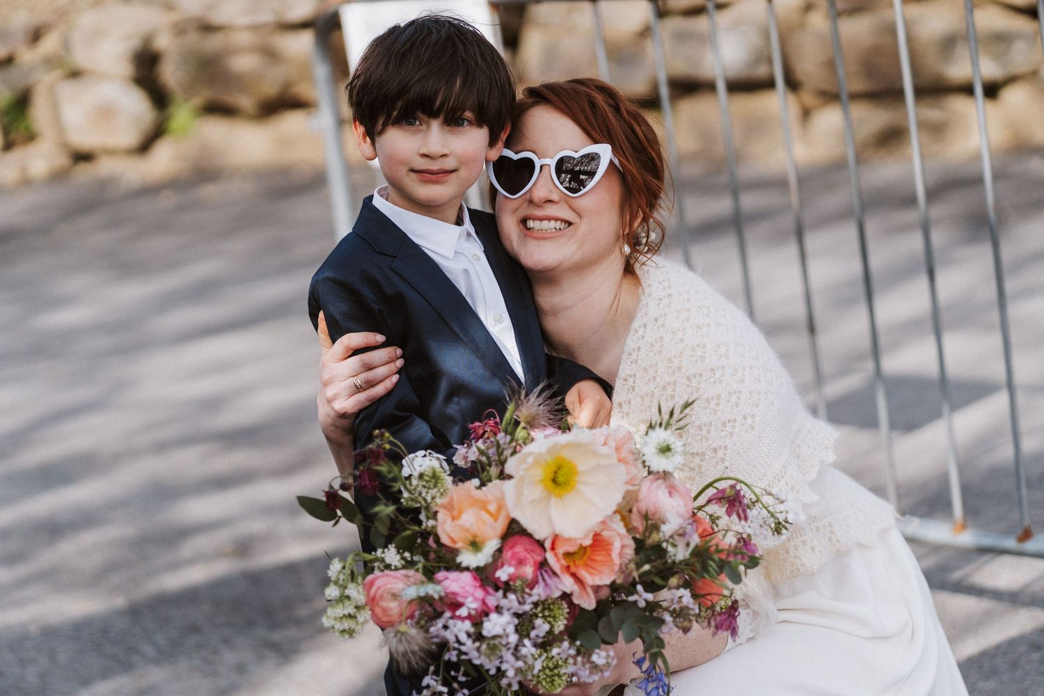 Bride in white lace dress with heart sunglasses hugs young boy in suit, holding colorful wedding bouquet outdoors.