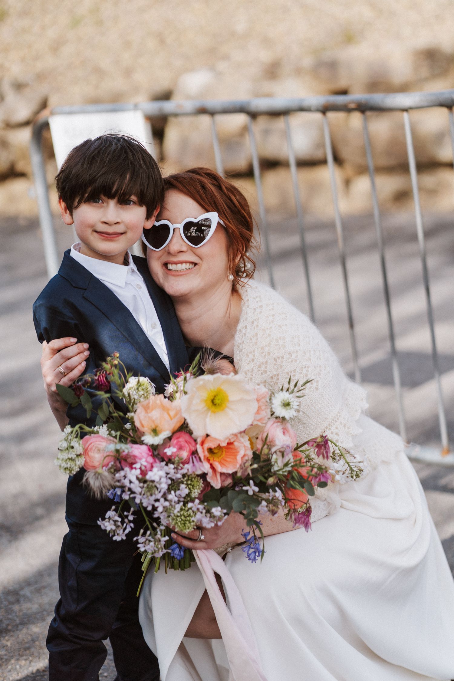 Bride in white dress with heart sunglasses hugs young boy in suit while holding colorful floral bouquet.