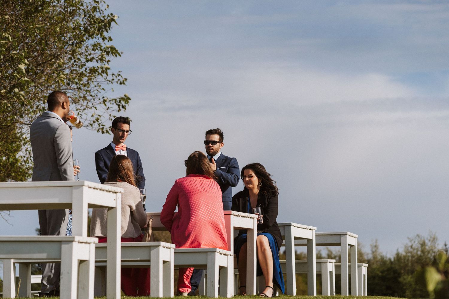 Elegantly dressed guests socializing outdoors at a wedding reception with white chairs under a blue sky.