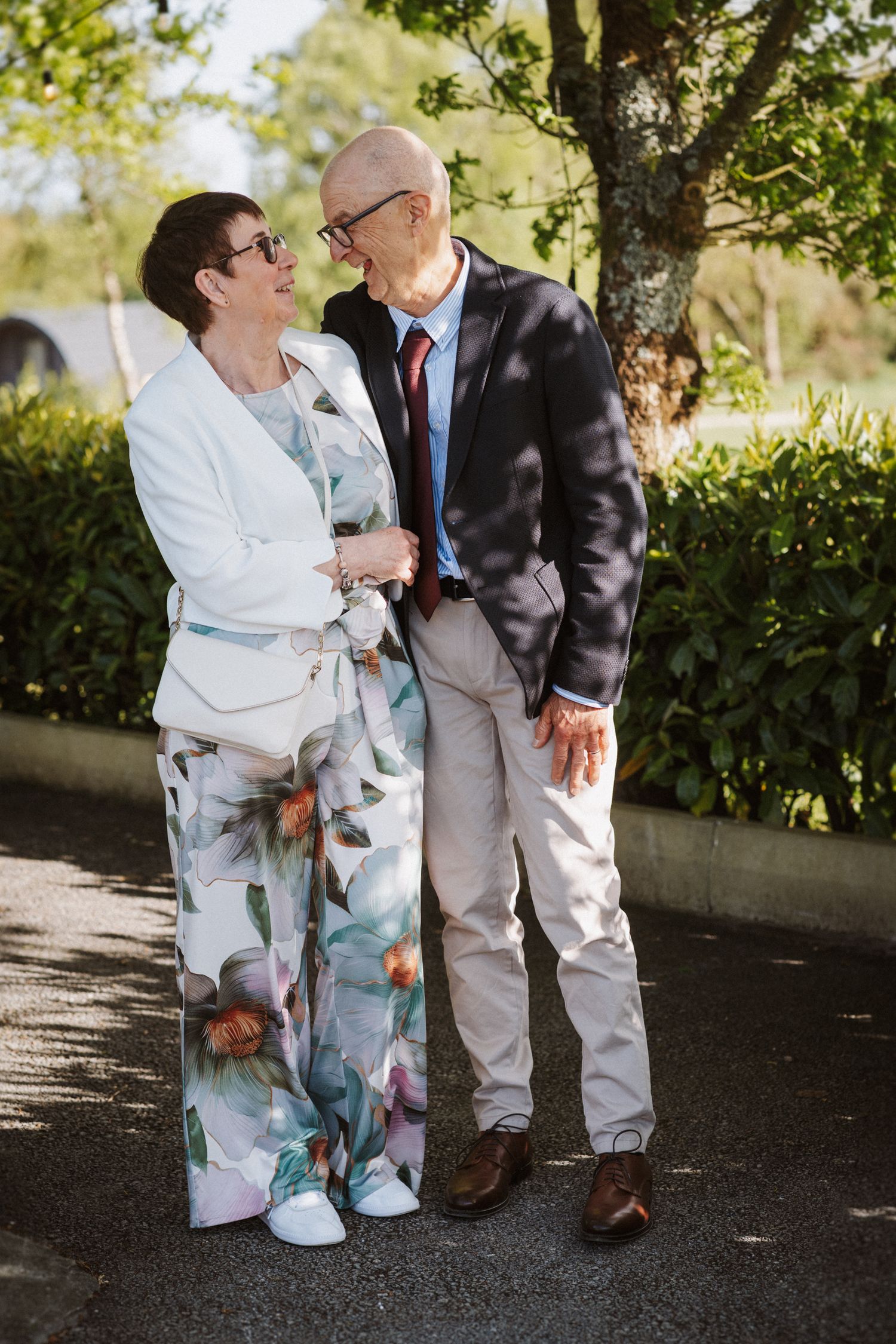 Elegant older couple in formal attire smile at each other outdoors surrounded by green foliage.