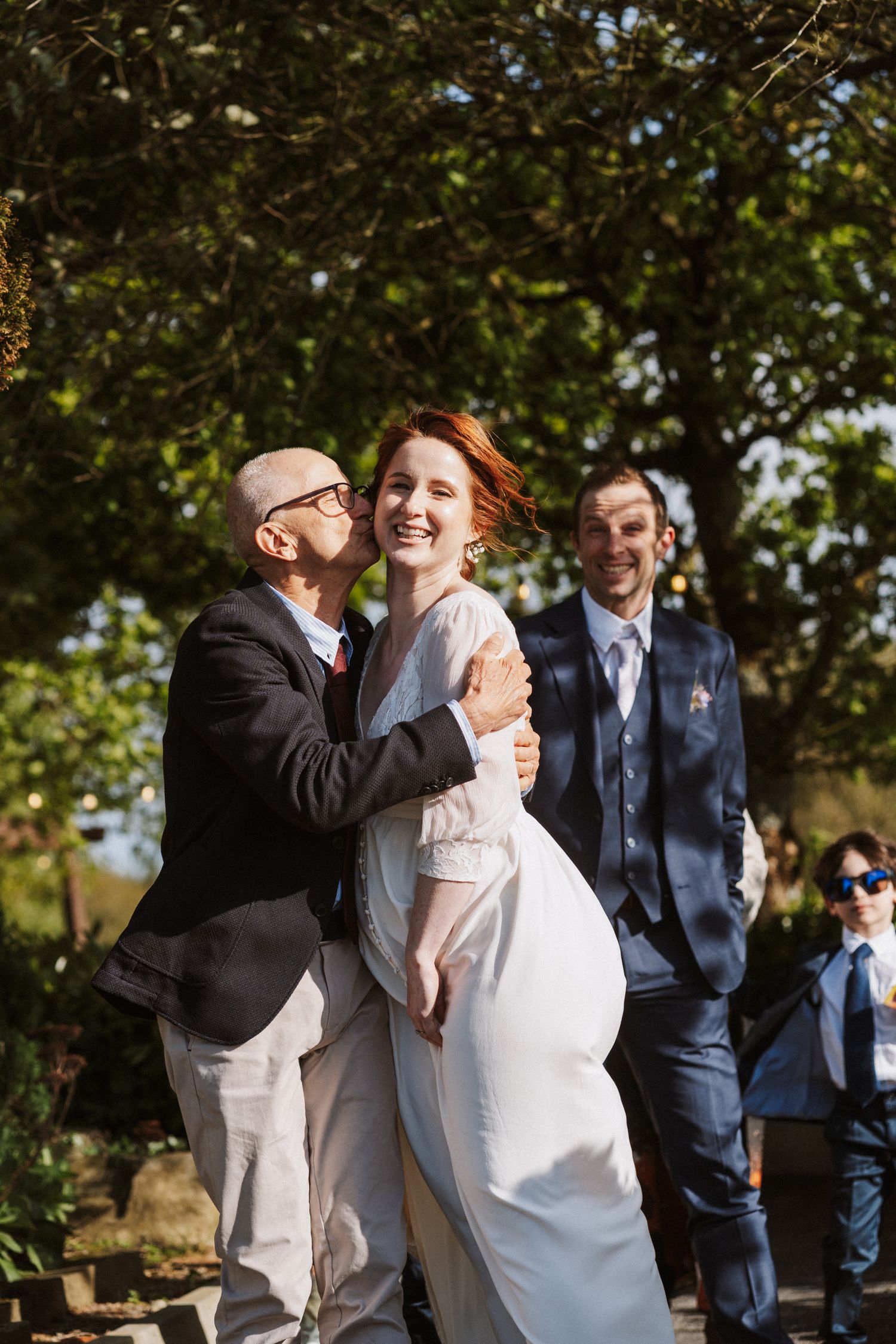 Groom kisses bride in white dress outdoors at wedding ceremony with smiling guests in background.