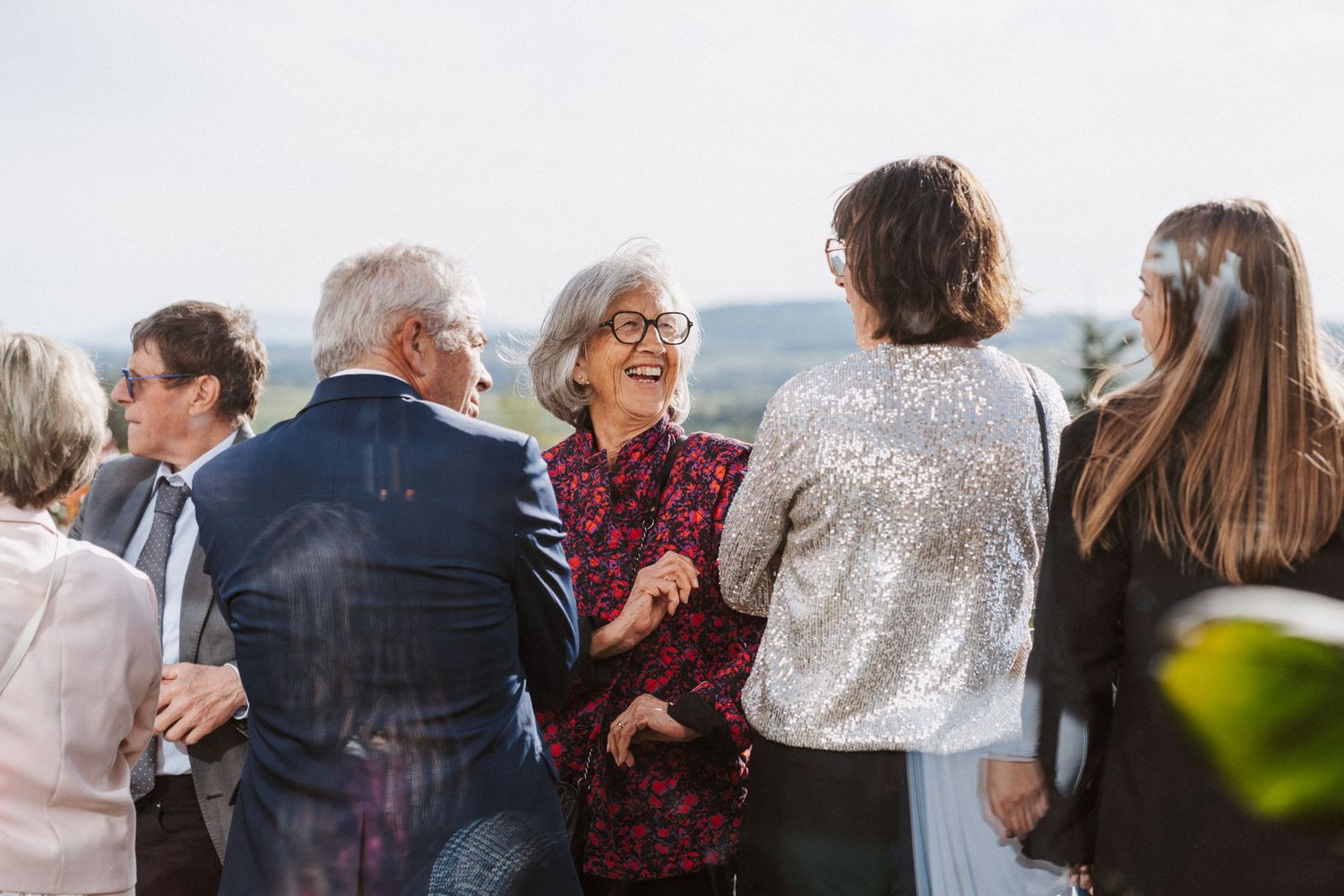 Elegantly dressed wedding guests mingle and laugh outdoors with scenic hills visible in the background.
