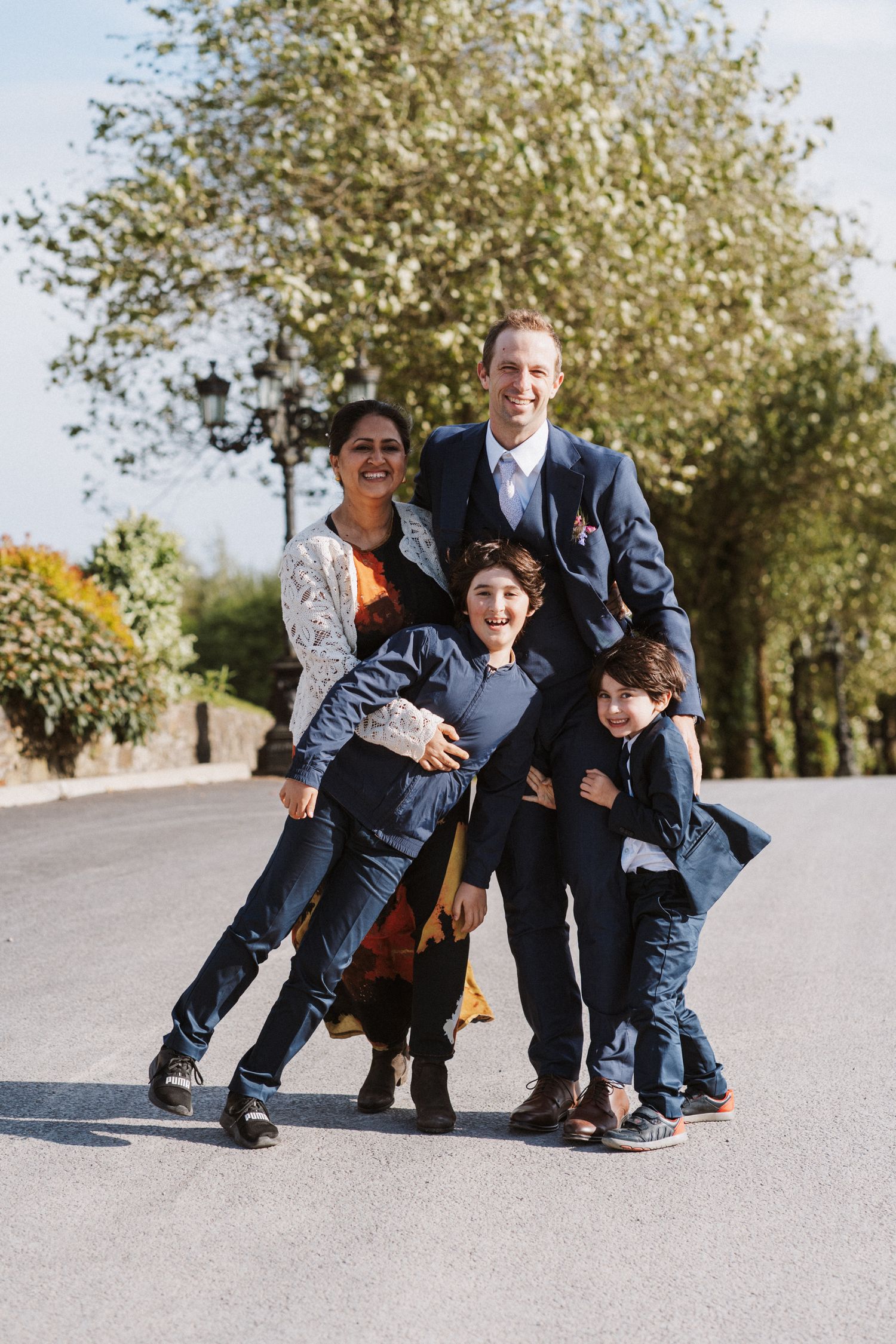 Happy family of four poses playfully on a tree-lined road during a wedding celebration outdoors.