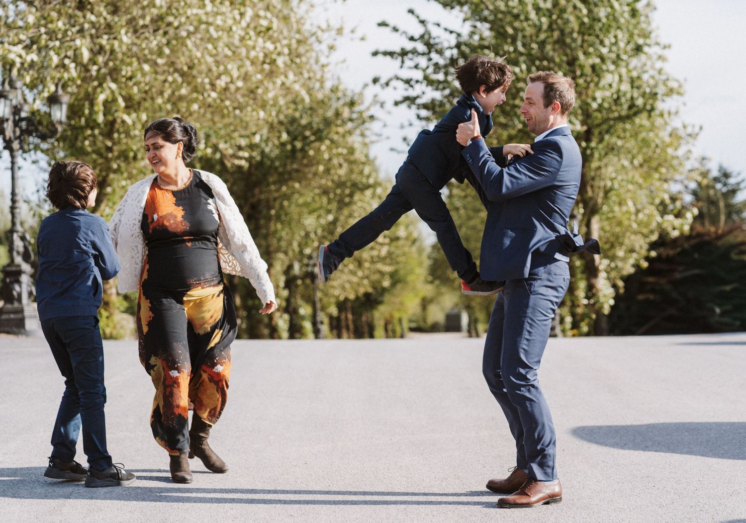 Man in blue suit playfully lifts a child while a woman and young boy watch outdoors at a wedding celebration.