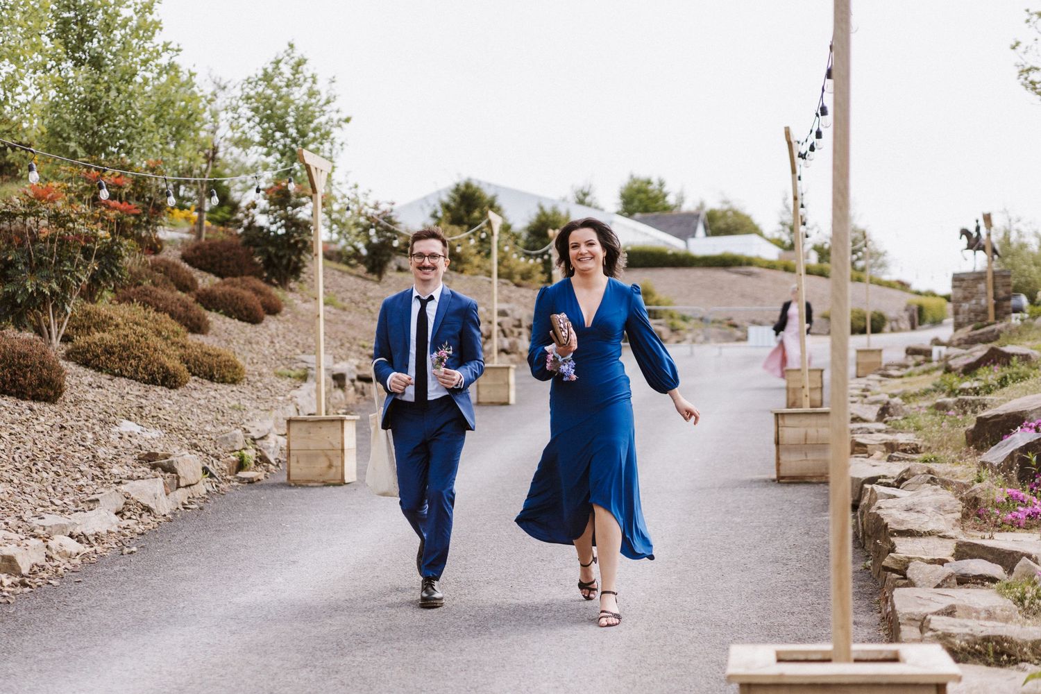 A couple in formal attire runs joyfully down a path at an outdoor wedding venue on a cloudy day.
