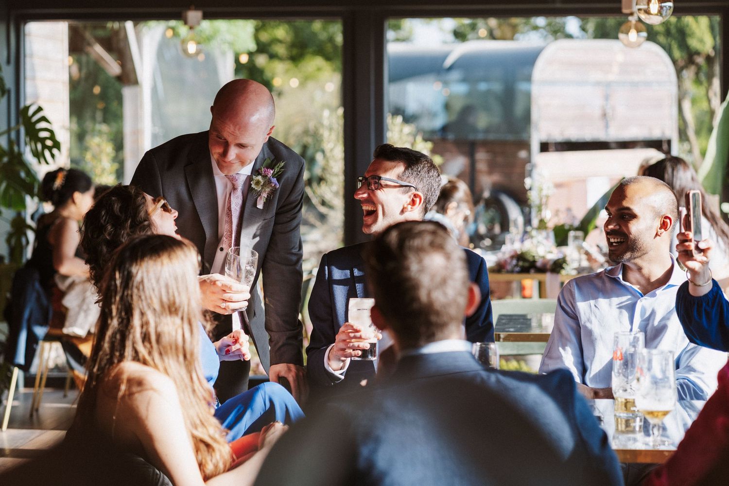 Bald groom gives a heartfelt wedding speech to smiling guests seated at tables in a warmly lit reception venue.
