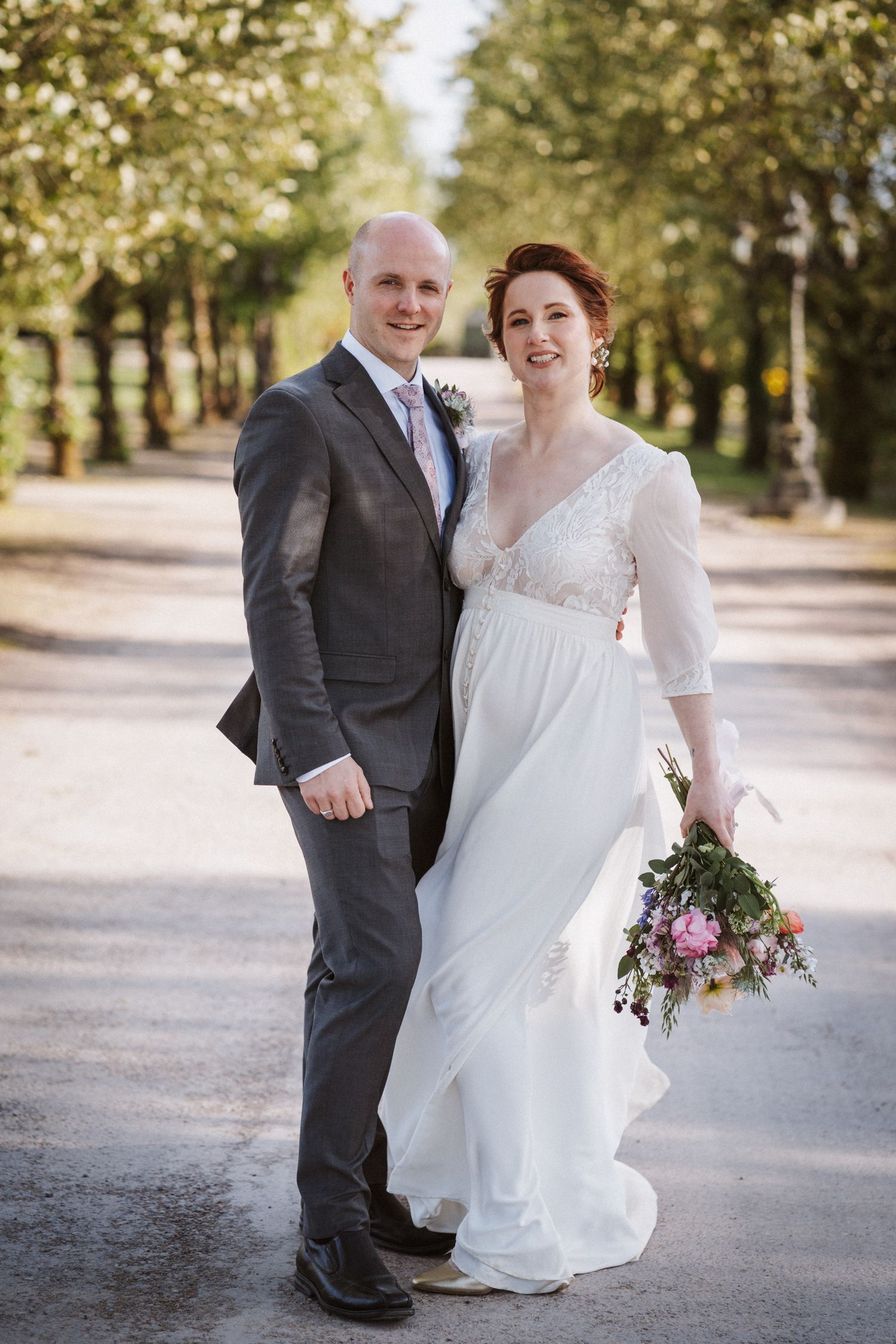 Bride in white lace gown holding wildflower bouquet poses with groom in grey suit on tree-lined path.