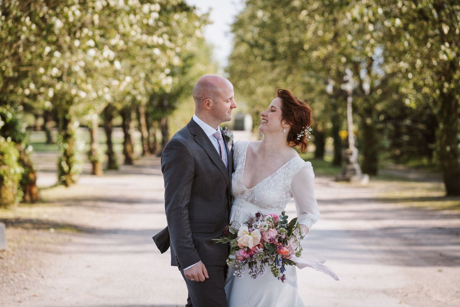 Bride and groom share a romantic moment on a tree-lined path, bride holding a colorful bouquet on their wedding day.