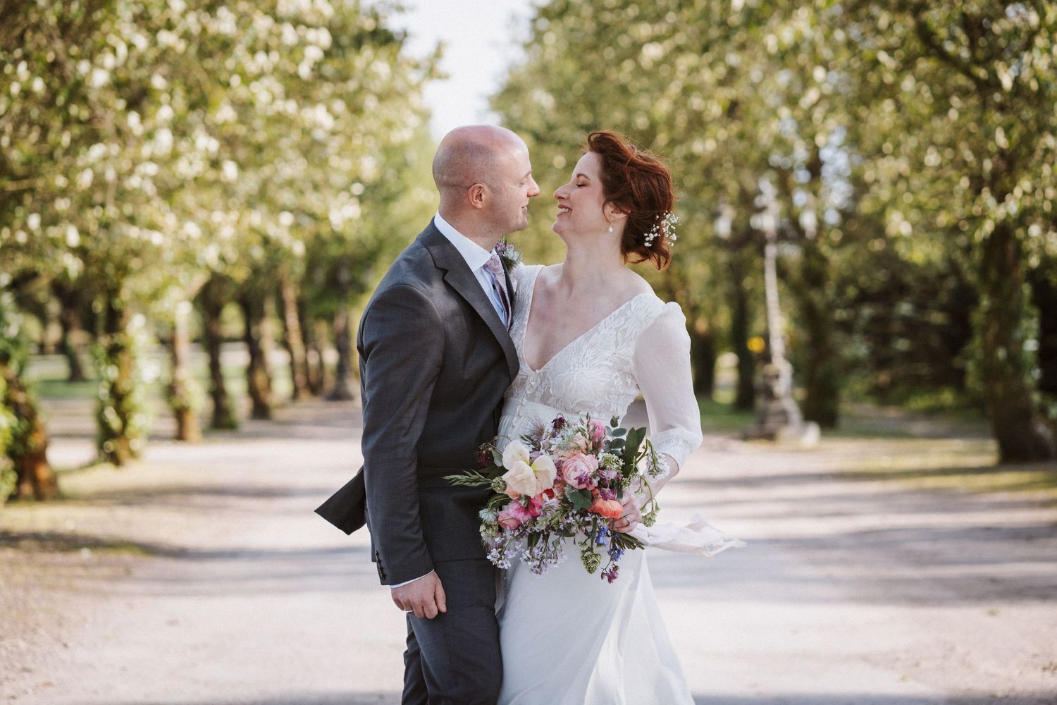 Bride and groom share a romantic moment on a tree-lined path, bride holding a colorful bouquet.