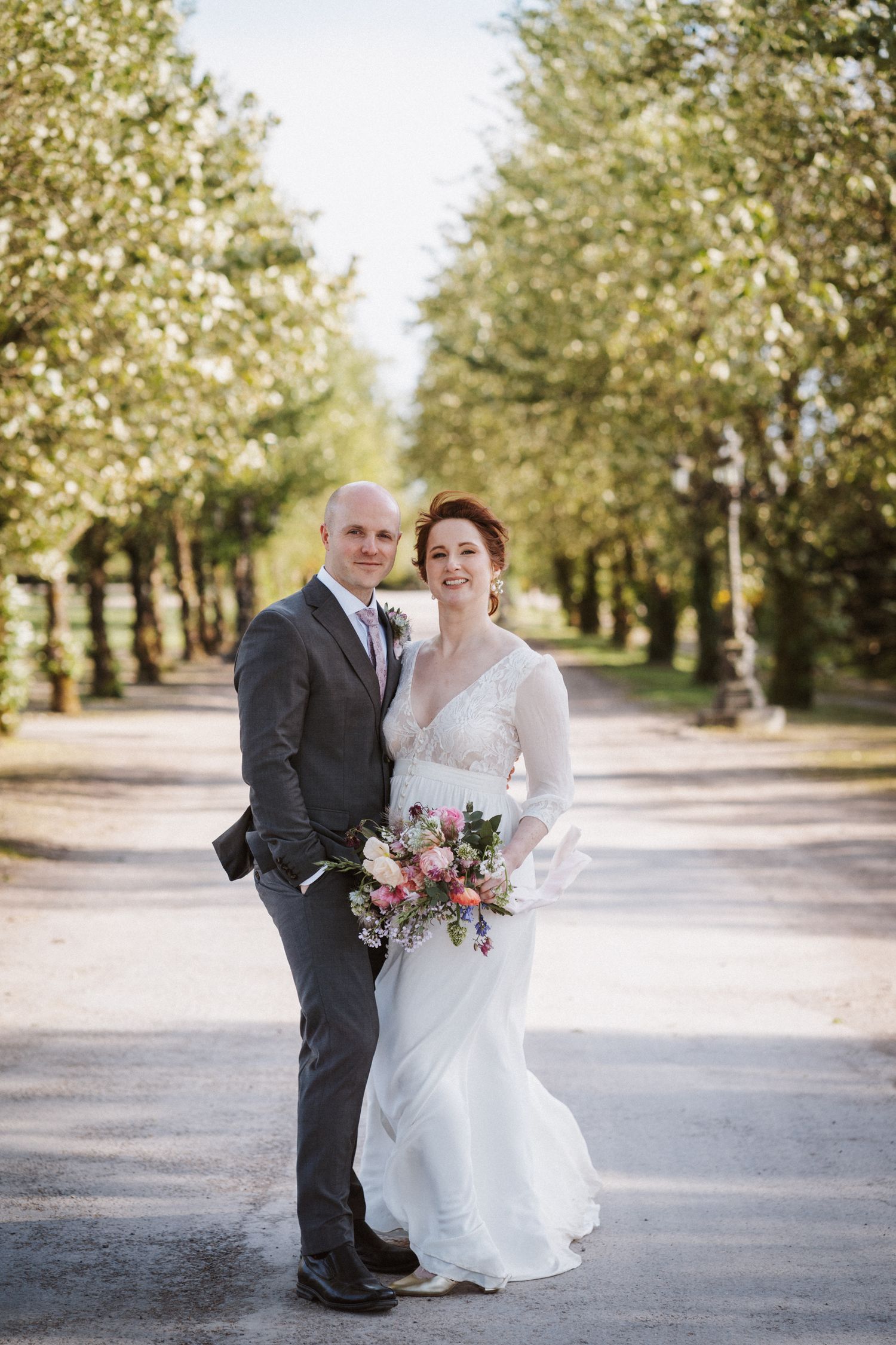 Bride and groom pose on tree-lined path, bride holding pink bouquet in lace wedding dress.