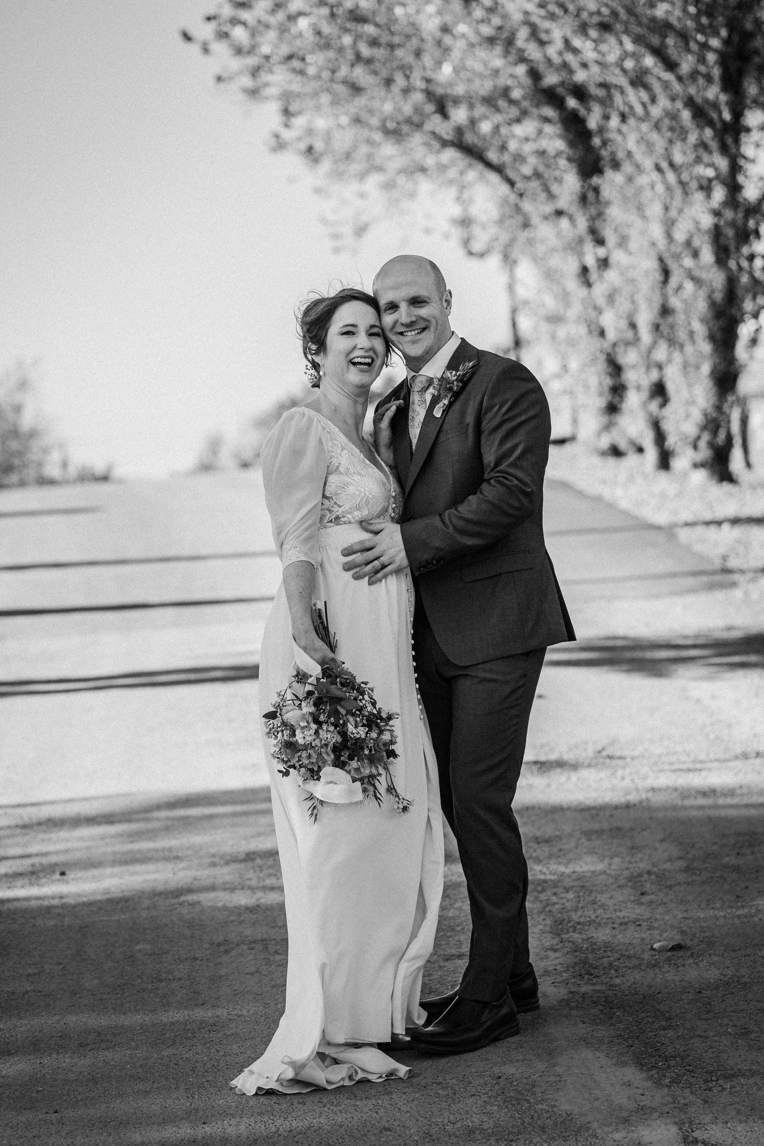 Black and white wedding photo of smiling bride holding bouquet and groom embracing outdoors near tree-lined path.
