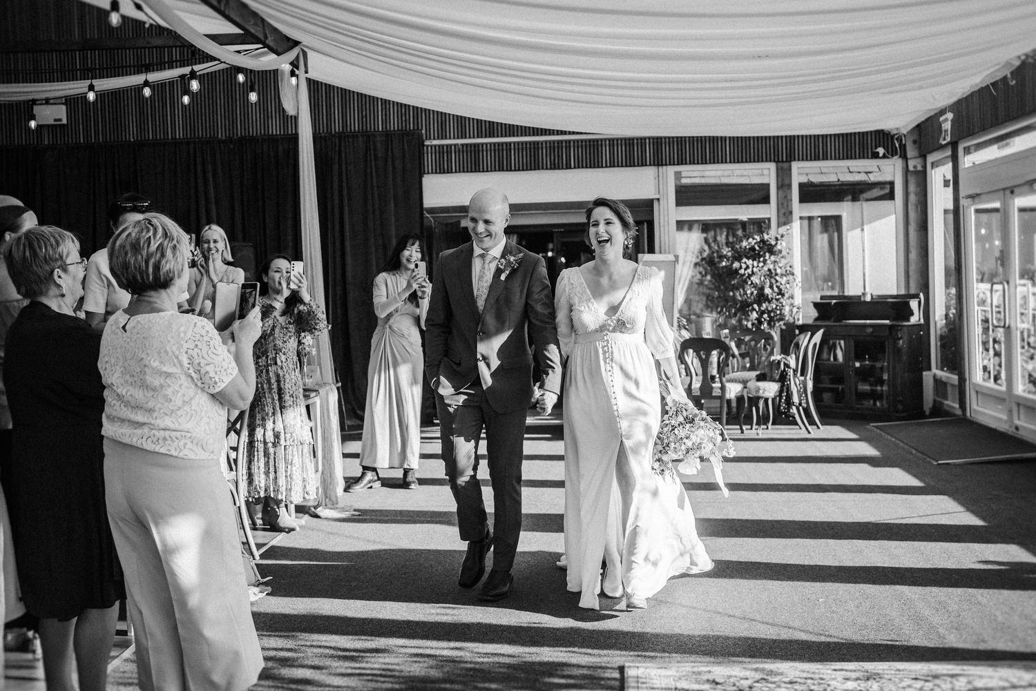 Black and white photo of bride and groom walking down aisle as guests applaud at outdoor wedding.