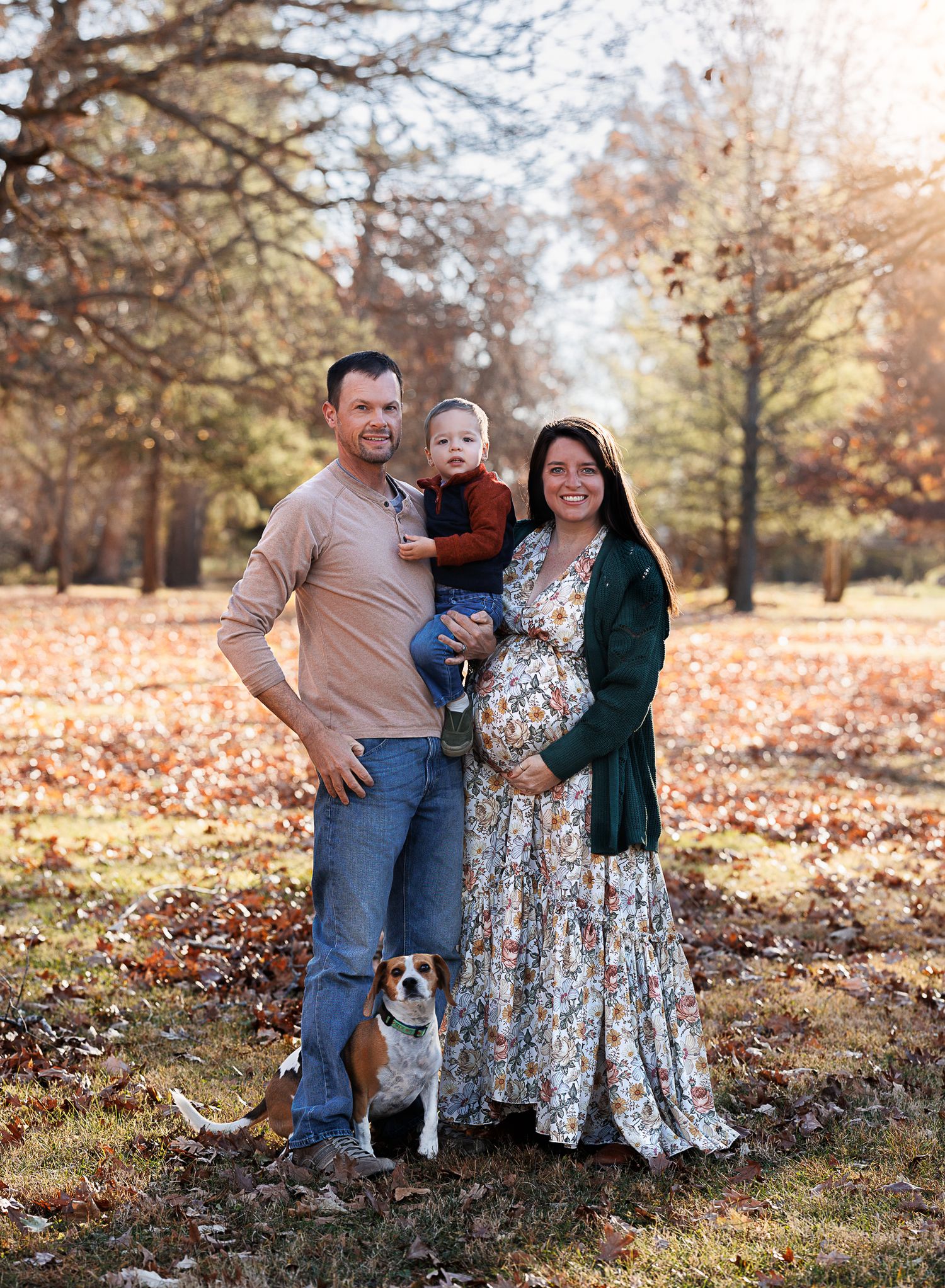 A pregnant woman in a floral dress poses with her husband, toddler son, and beagle dog in an autumn park.