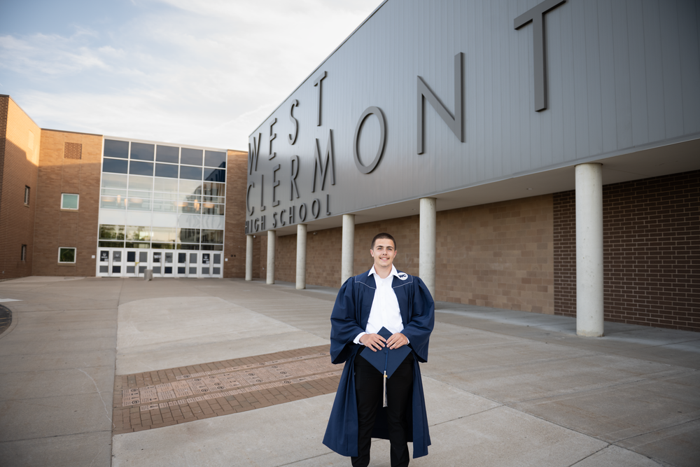 Graduate in blue cap and gown stands proudly outside West Clermont High School building.