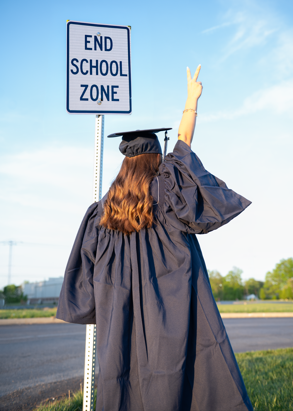 Graduate in cap and gown giving peace sign next to End School Zone street sign, celebrating graduation outdoors.