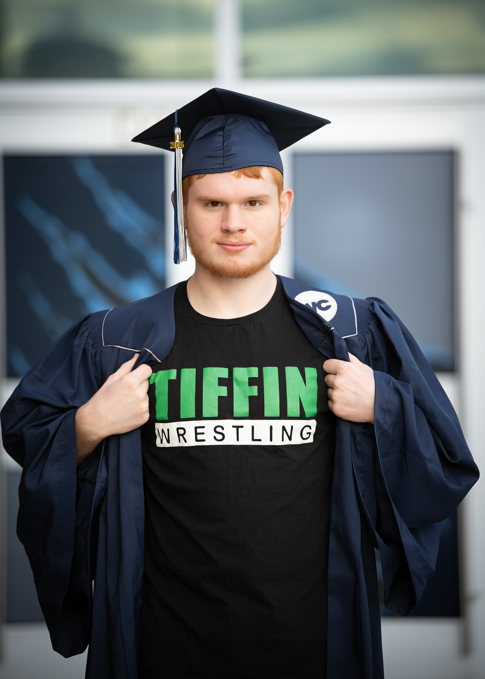 Young man in graduation cap and gown proudly displaying Tiffin Wrestling t-shirt outdoors.