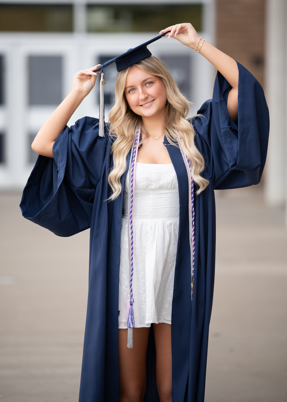 Smiling graduate in navy cap and gown with honor cords poses outdoors for senior portrait photo.
