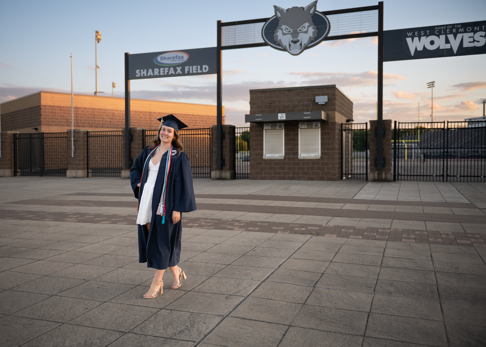 Graduate in cap and gown poses at Sharifax Field entrance with Wolves mascot sign at sunset.