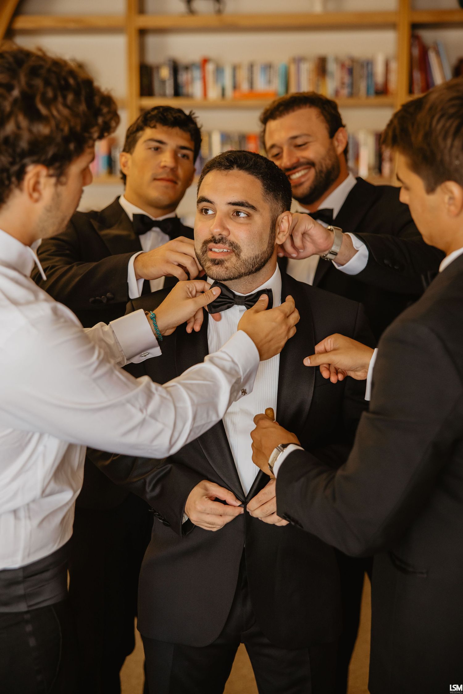 Groomsmen helping the groom adjust his bow tie and tuxedo jacket before the wedding ceremony.