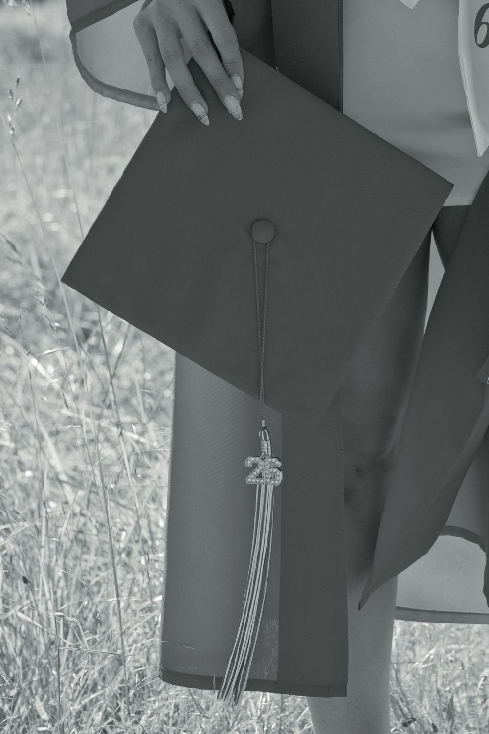 Graduate holding a mortarboard cap with class of 25 tassel in a monochrome outdoor photo.