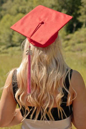 Blonde woman wearing a red graduation cap with tassel and 26 charm, viewed from behind with green nature background.