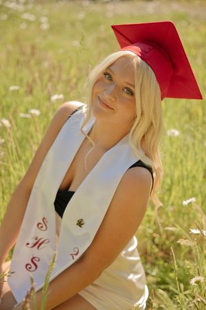 Smiling blonde graduate in red cap and white sash sits in a sunny wildflower field for senior portrait.