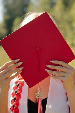 Graduate holds red mortarboard cap with 2021 tassel charm, wearing white stole with red accents outdoors.