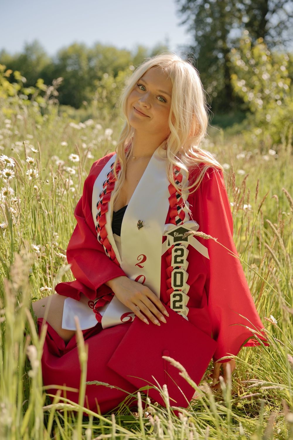 Blonde woman in red graduation gown and 2022 sash sitting in a wildflower meadow on a sunny day.