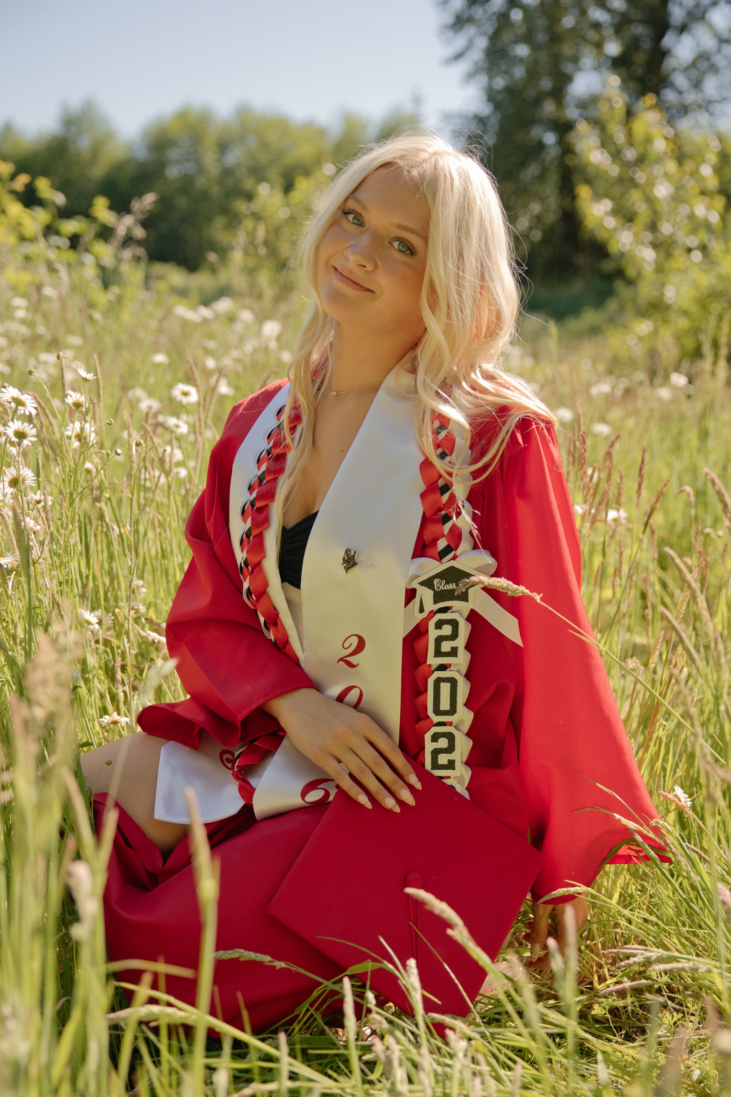 Blonde woman in red graduation gown and 2022 sash sitting in a wildflower meadow on a sunny day.