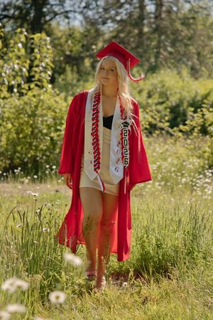 Blonde woman in red graduation cap and gown standing in a green meadow with wildflowers on a sunny day.