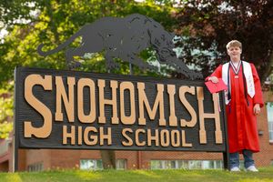 Graduate in red cap and gown poses by Snohomish High School sign on sunny day.