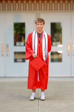 A smiling graduate in a red cap and gown holds their cap while standing outside school doors with honor cords.