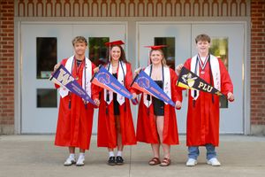 Four high school graduates in red caps and gowns hold college pennants outside a brick building.