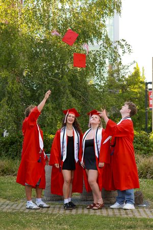 Four graduates in red caps and gowns toss their mortarboards in the air outdoors near green trees.