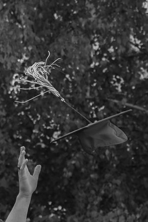 Black and white photo of a hand tossing a graduation cap with flowers into the air against a tree backdrop.