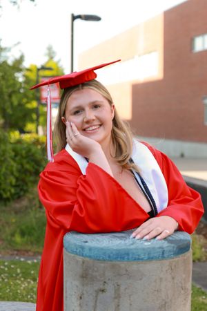 Smiling female graduate in red cap and gown posing outdoors leaning on concrete pillar.
