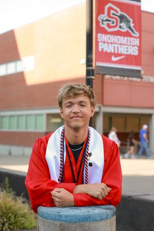 A smiling male graduate in red gown with white stole poses in front of Snohomish Panthers school sign.