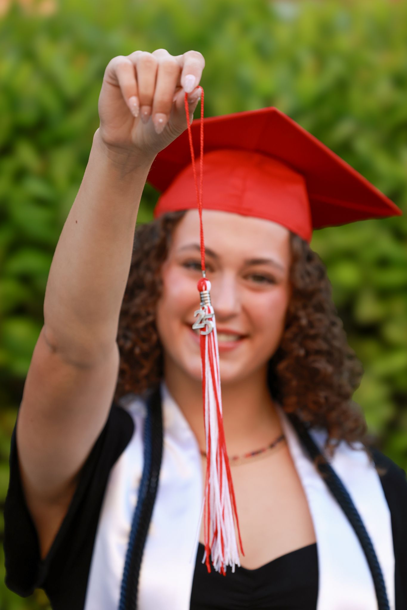 A smiling young woman in a red graduation cap and gown holds up her tassel outdoors.