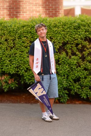 High school senior boy poses outdoors wearing graduation sash and holding University of Washington pennant.