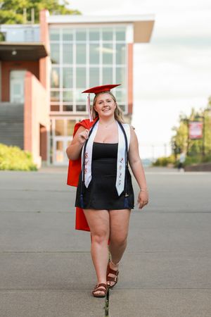 Smiling female graduate in red cap and gown walks confidently outside a brick university building on graduation day.