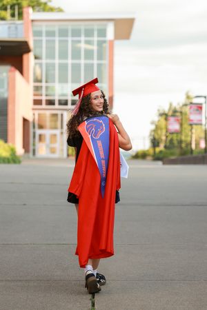 A smiling graduate in red cap and gown with Boise State sash walks outside a modern building.