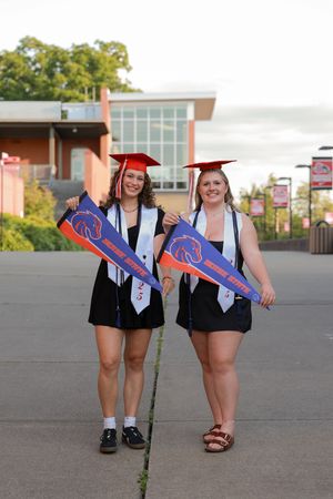 Two female graduates in red caps and gowns holding Boise State pennants outdoors.