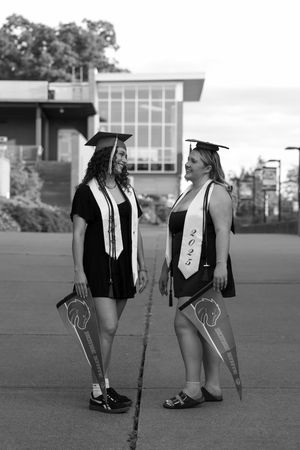 Two female graduates in black caps and white stoles smile at each other holding Boise State pennants in black and white.
