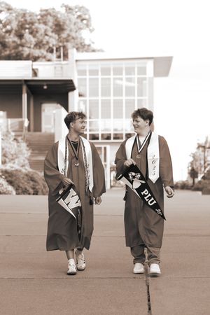 Two graduates in caps and gowns walk together holding college pennants for UW and PLU.