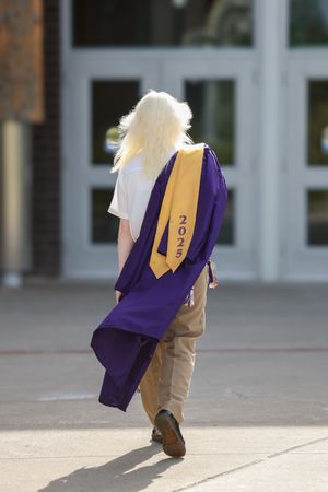Student walking away in purple graduation gown with gold 2025 sash, long blonde hair, approaching school entrance.