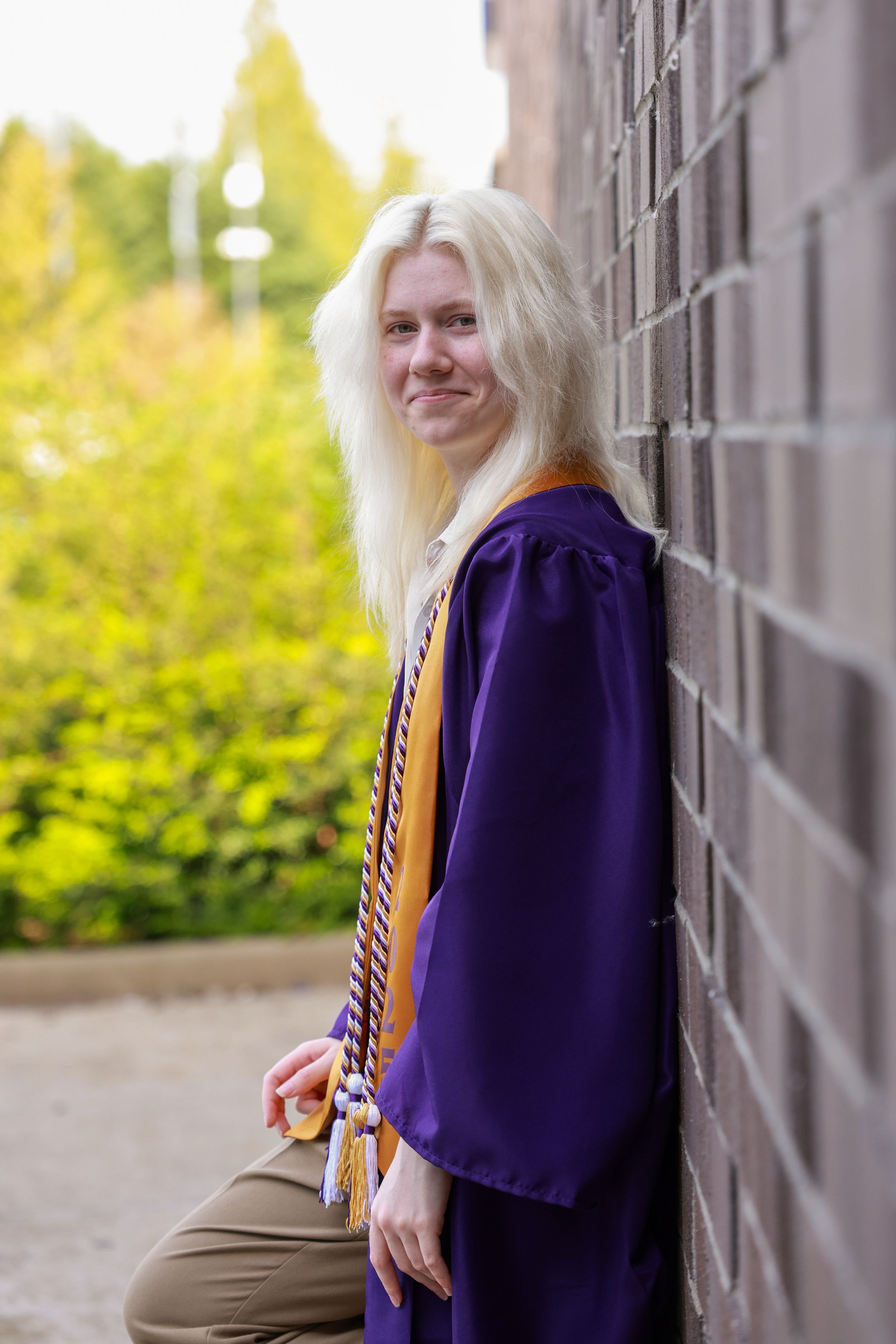 Smiling blonde graduate in purple cap and gown leaning against brick wall outdoors.