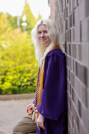 Smiling blonde graduate in purple cap and gown leaning against brick wall outdoors.