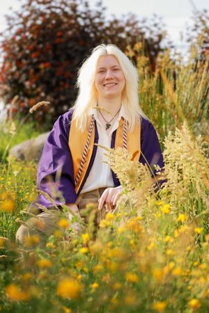 Smiling blonde graduate in purple and gold regalia sitting among wildflowers and tall grass outdoors.