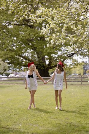 Two female graduates in white dresses and red caps hold hands walking across green grass beneath a flowering tree.