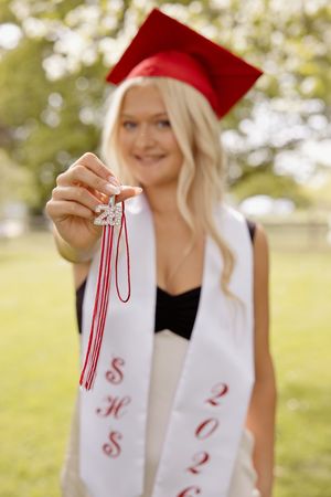 Blonde graduate in red cap holds up a sparkling 2026 tassel charm while wearing a white SHS stole.